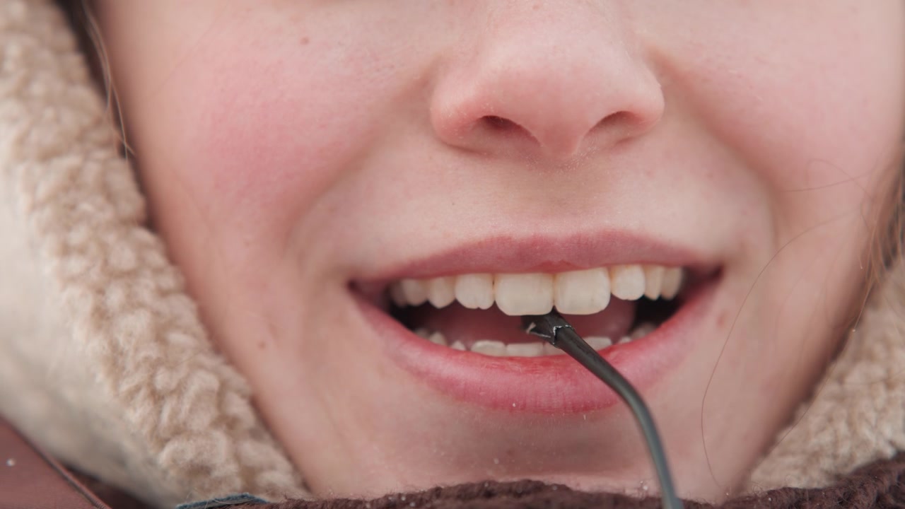 Tight frontal close-up of lower face with fleece hood and scarf, mouth open as thin black cord rests between lips and teeth; visible action shows careful adjustment in cold outdoor light