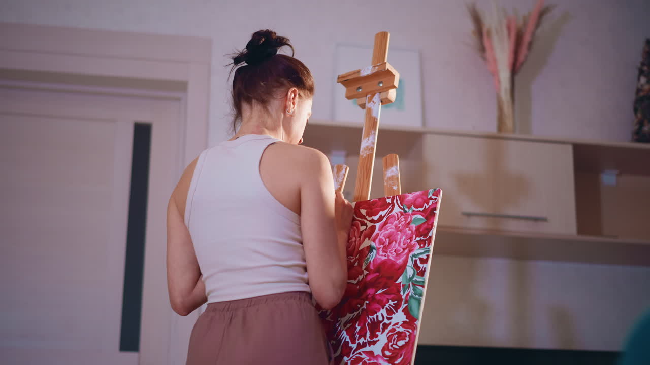 Young White Woman Painting Floral Canvas From Behind In Home Studio, Commission Piece With Bold Red And Pink Motifs, Textured Strokes, Fabric Pattern Influence And Calm Domestic Interior