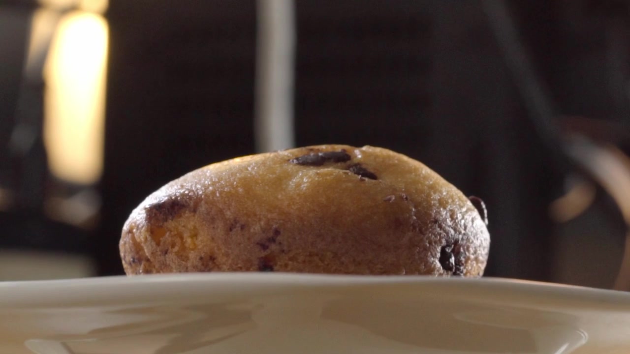 Primo piano macro di un muffin con gocce di cioccolato appena sfornato su piatto bianco con illuminazione soffusa sullo sfondo. Dessert da forno invitante che evidenzia la consistenza dorata e i pezzi di cioccolato.