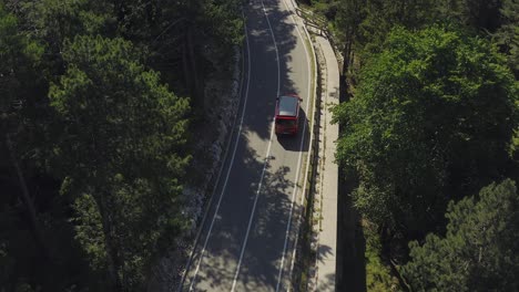 Red-minivan-with-black-roof-driving-along-winding-mountain-road-through-forest-in-Europe