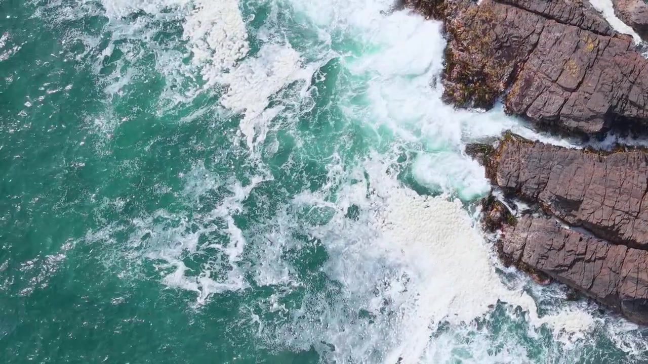 Vista aerea statica dall'alto, cinematografica, di potenti onde oceaniche turchesi e schiuma bianca che si infrangono contro aspre scogliere rocciose sulla costa selvaggia dell'isola di Chiloé nella Patagonia cilena.