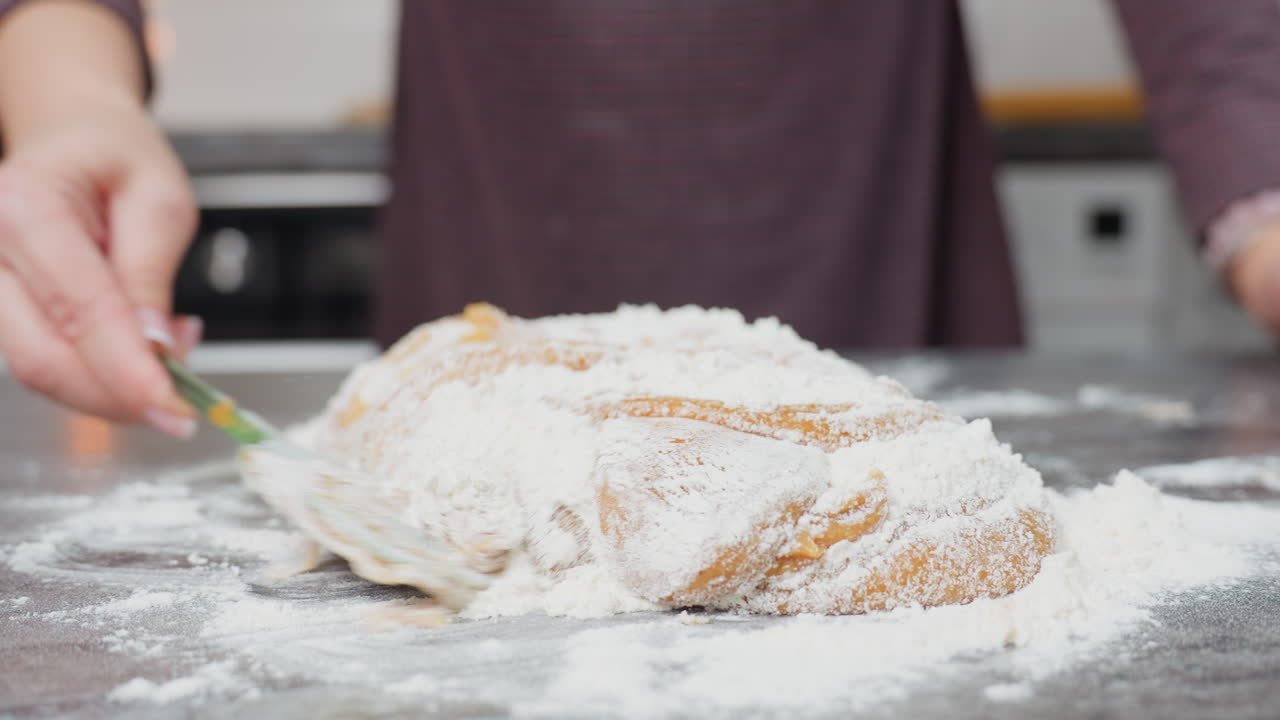 Close-up of baker in maroon cloth mixing soft dough with flour on countertop, hands kneading, folding, and blending ingredients for baking
