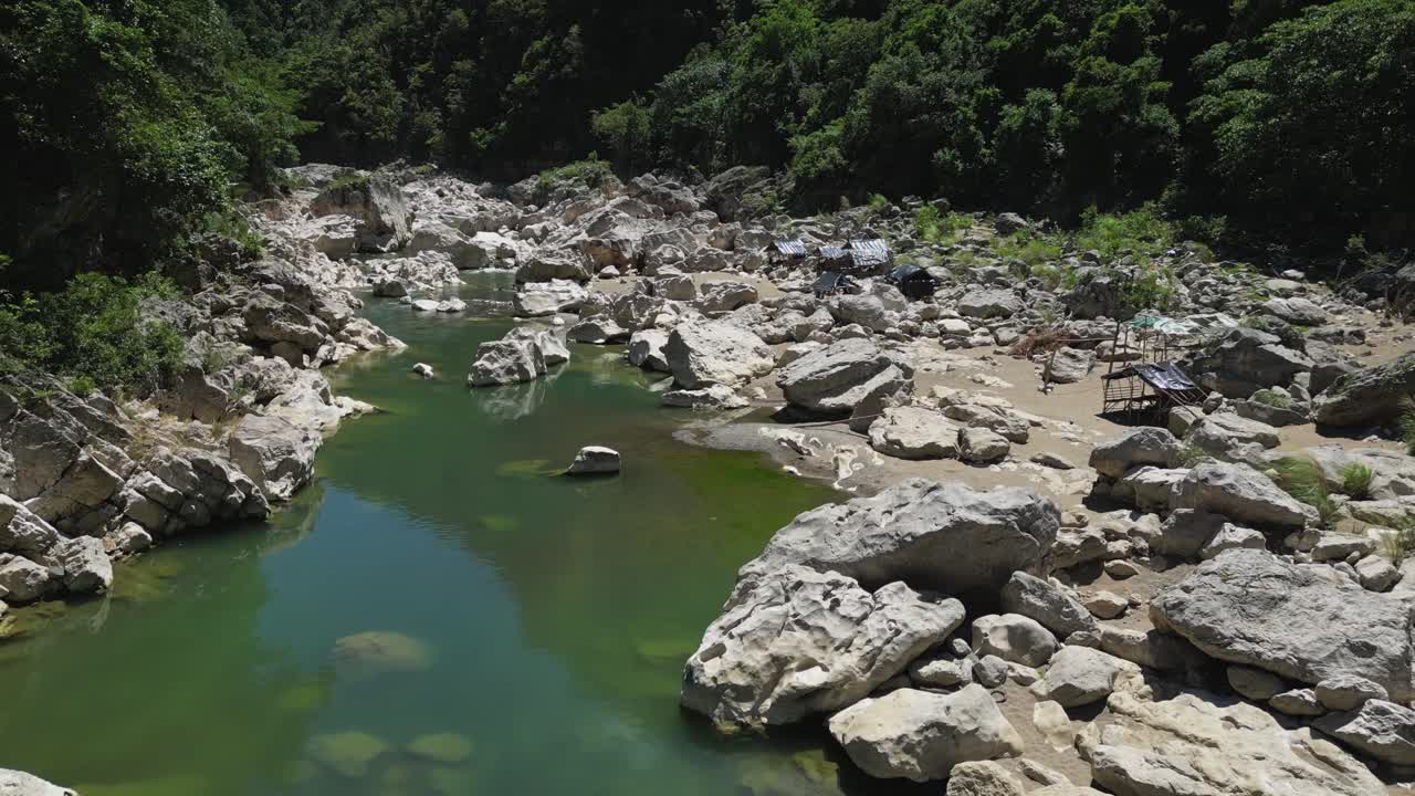 Forward shot focuses on the river and small rocks in the middle, with cottages lining the edges beside Tinipak River, Philippines