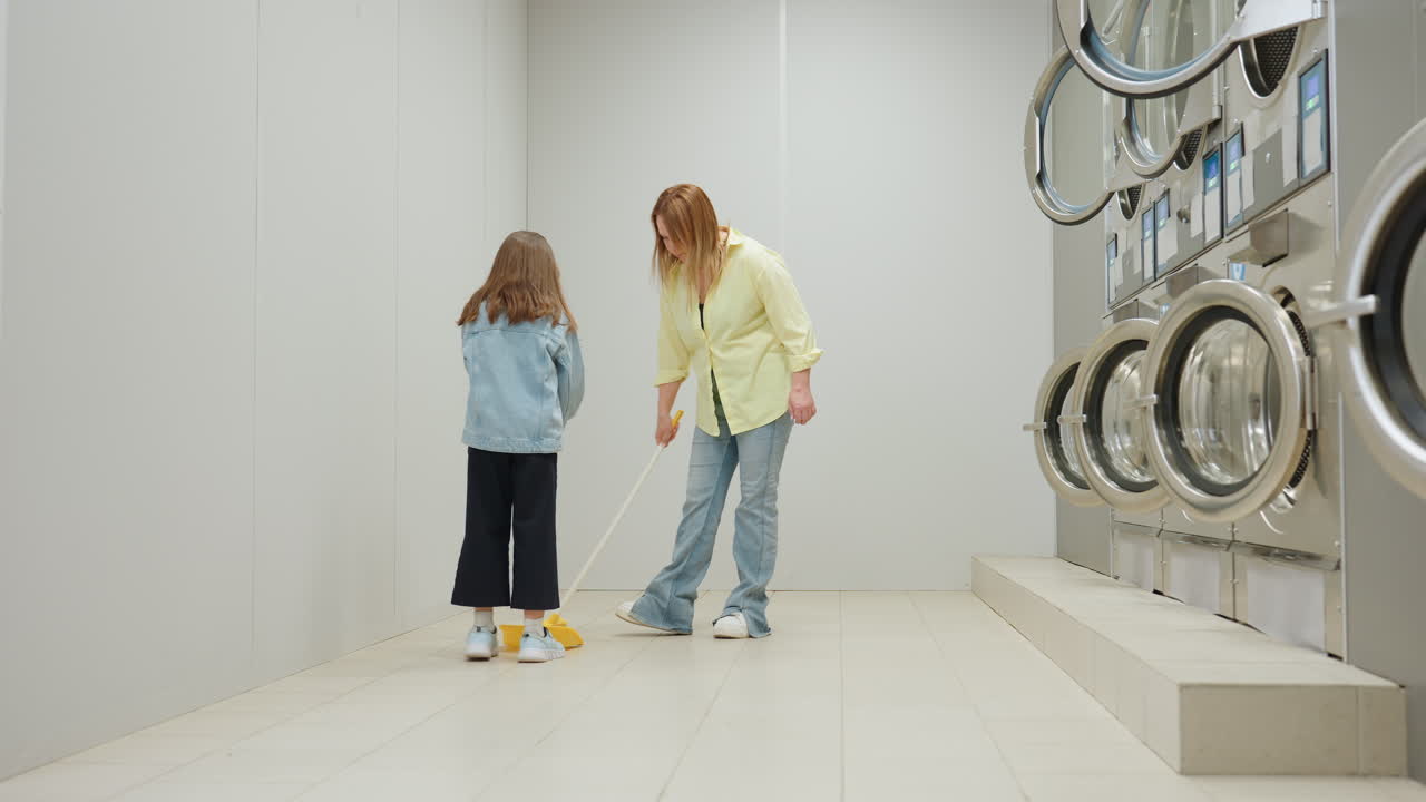 Long shot of mother sweeping tiled floor while daughter holds dustpan in bright laundromat, teamwork cleaning near stainless industrial washers, guidance, hygiene, cooperation between machines