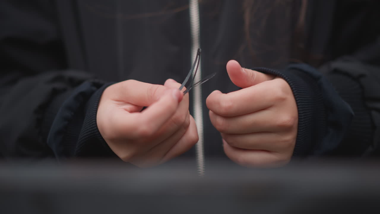 Closeup hands untangling earbud cable during drizzle, asian commuter in dark jacket patiently frees tiny plug and inspects connection, shallow depth of field with soft overcast light, everyday ritual