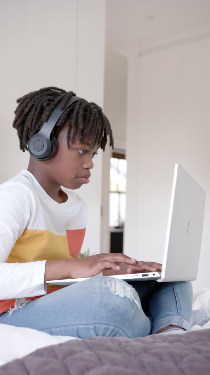 Vertical video of happy african american boy sitting on bed using laptop at home, slow motion