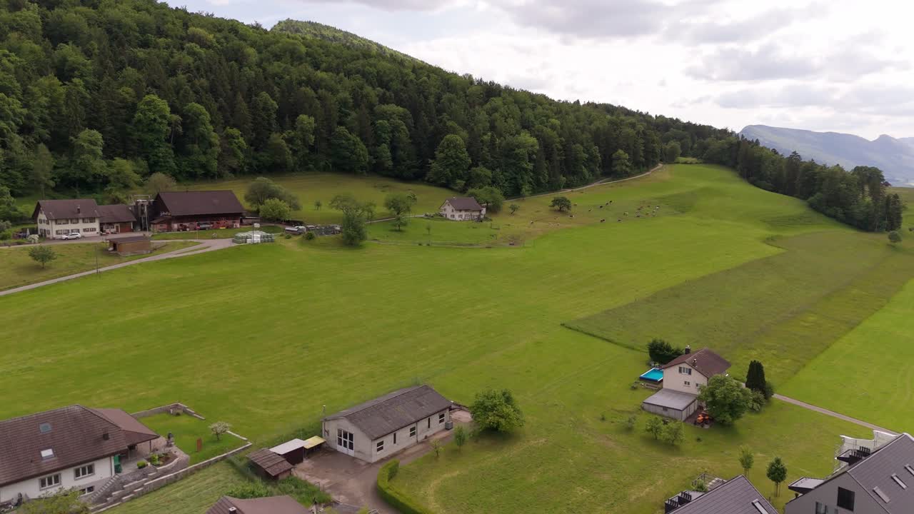 Modern multi.family units and traditional swiss houses on top of green mountains. Sunny day in Switzerland. Green pasture fields in alps. Aerial forward wide shot.