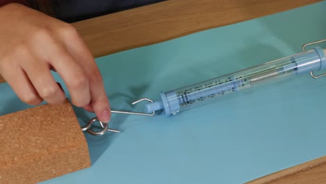 Hands manipulate a spring scale to measure force on a cork block with weights, illustrating friction principles