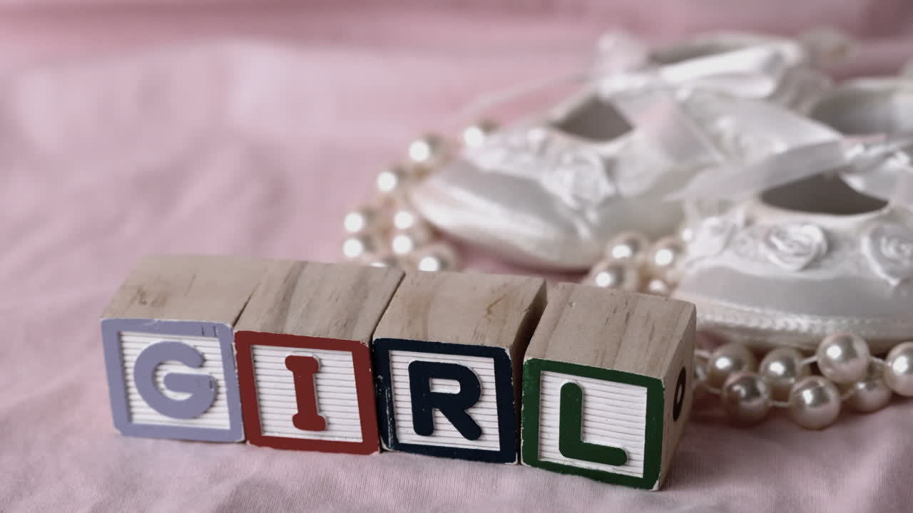Girl in letter blocks beside booties and pearls on pink blanket