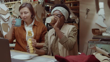 women drinking coffee, eating pastry and using laptop in workshop