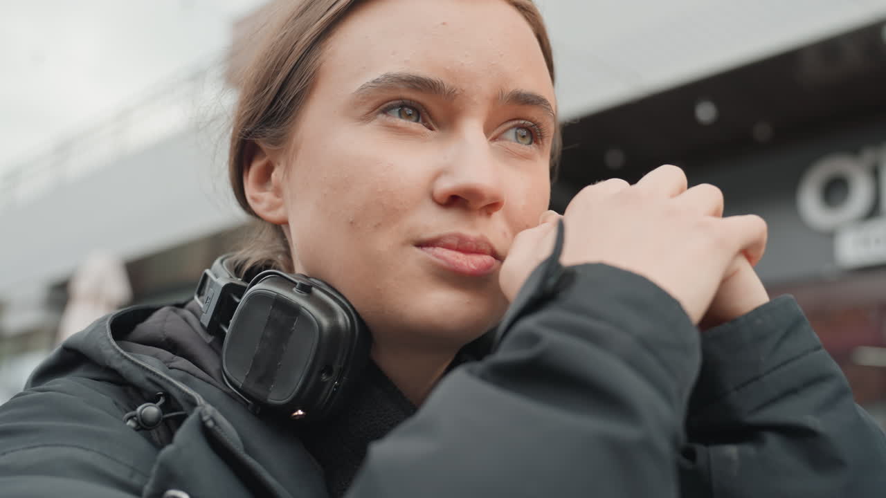 Белая женщина в наушниках стоит на улице, сцепив руки, смотрит вдаль с задумчивым выражением лица. Зимнее пальто, легкий снег, городской фон с витринами магазинов, откровенный крупный план портрета