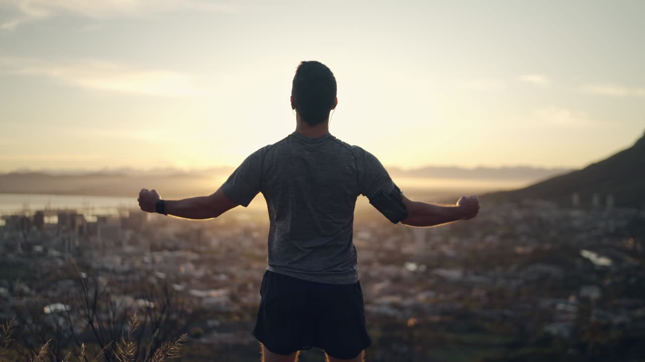 vista trasera de un atleta masculino saludable y deportivo estirando las manos con los puños cerrados celebrando su éxito frente al sol naciente en la cima de la montaña: hombre motivado celebrando el logro de sus objetivos