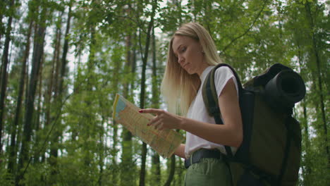 young caucasian woman looking for direction on a map while hiking in the forest. happy girl while hiking in nature and orienteering with help of a map