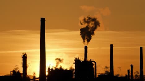 a striking sunset silhouette of industrial structures, featuring smoke stacks emitting fumes against a beautiful orange and yellow sky, capturing the essence of urban industry