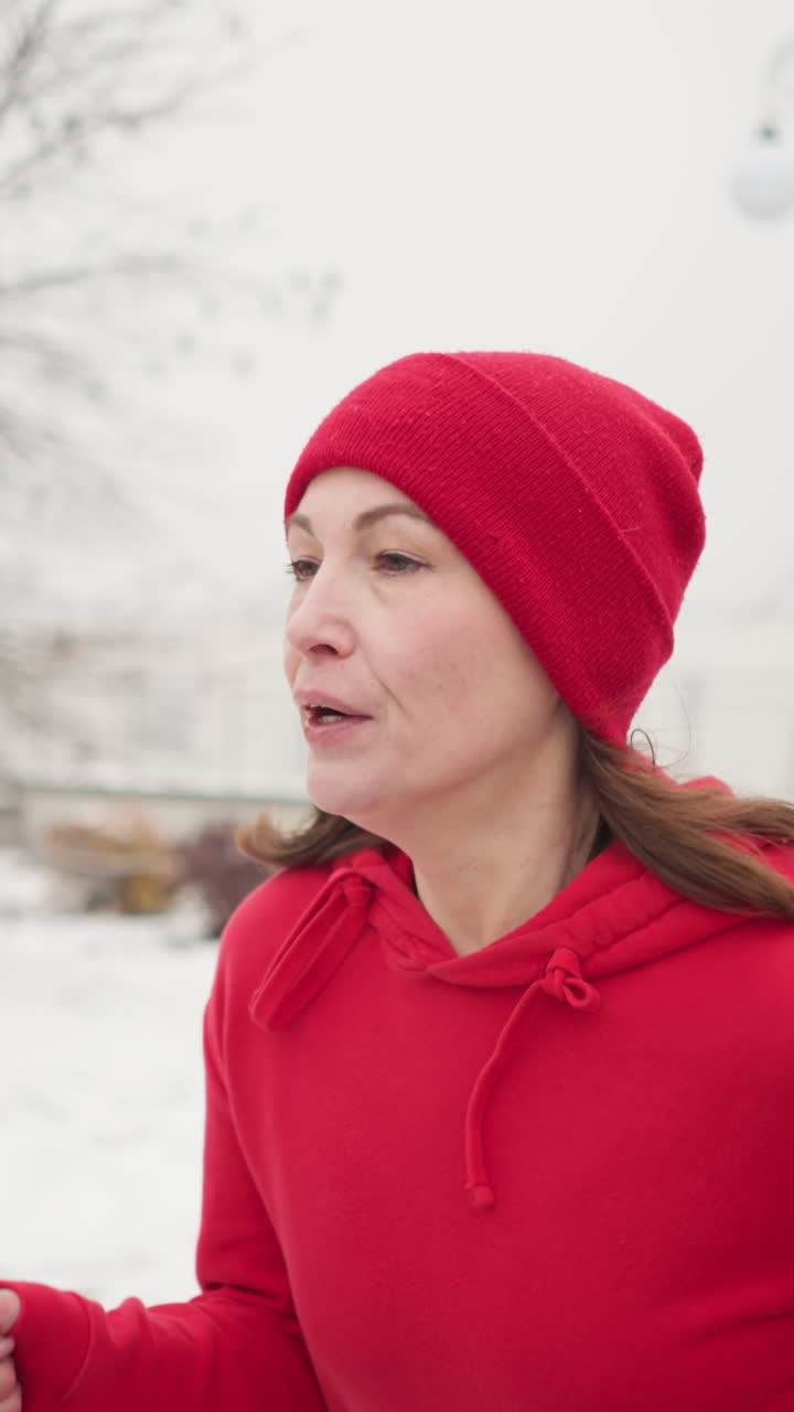 close-up of woman wearing red beanie and hoodie jogging outdoors on snowy winter morning surrounded by serene park trees with light posts and snow covered benches in blurred background