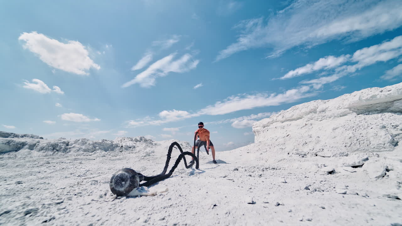 Shirtless man during working out with battle ropes in nature. Young muscular man training with ropes on the mountain against the blue sky. High intensive workout.