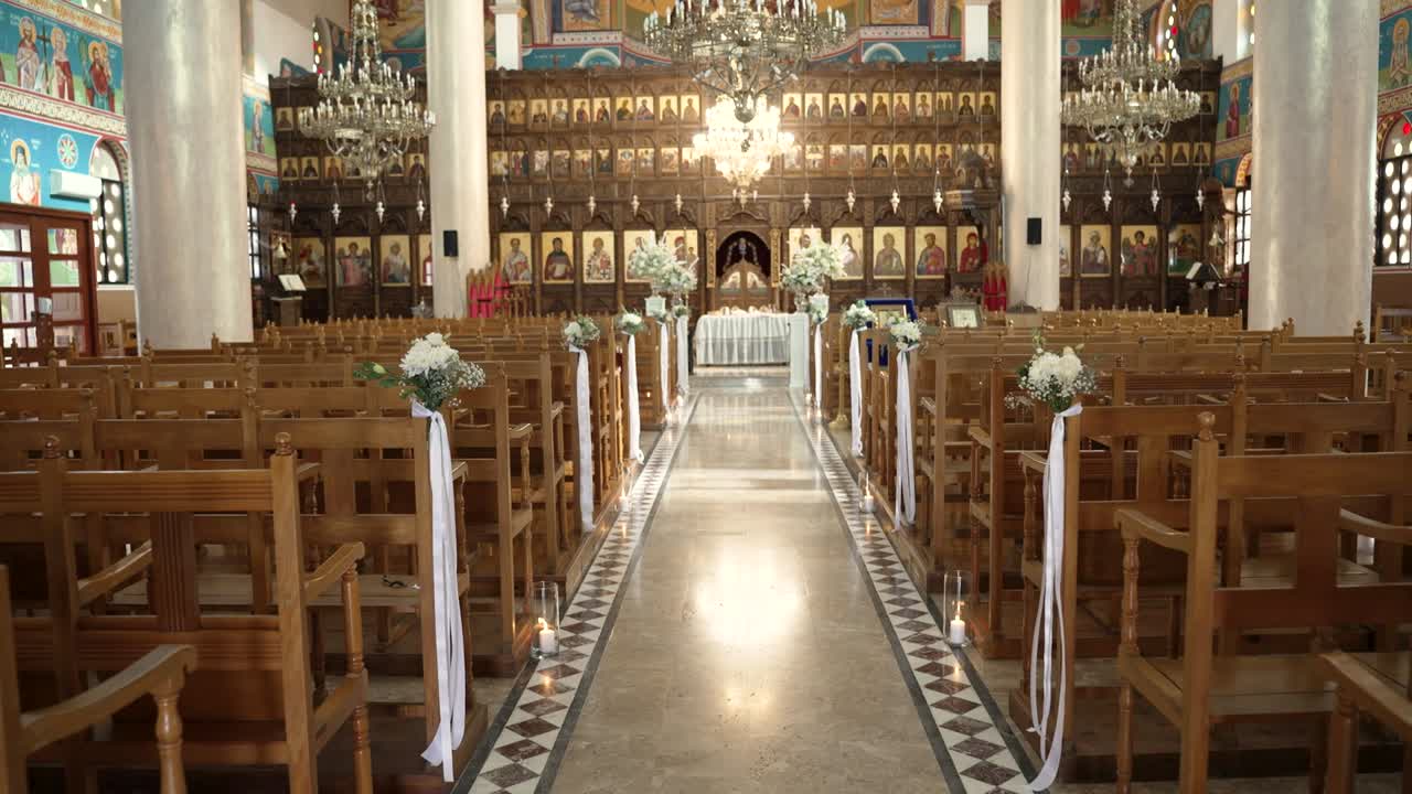 File di sedie di legno riempiono la chiesa, conducendo a un altare decorato. Composizioni floreali e nastri bianchi aggiungono dettagli alla navata. Gli invitati si raduneranno presto per la cerimonia.