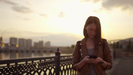 linda chica de jengibre usando teléfono móvil mientras camina por el terraplén al atardecer