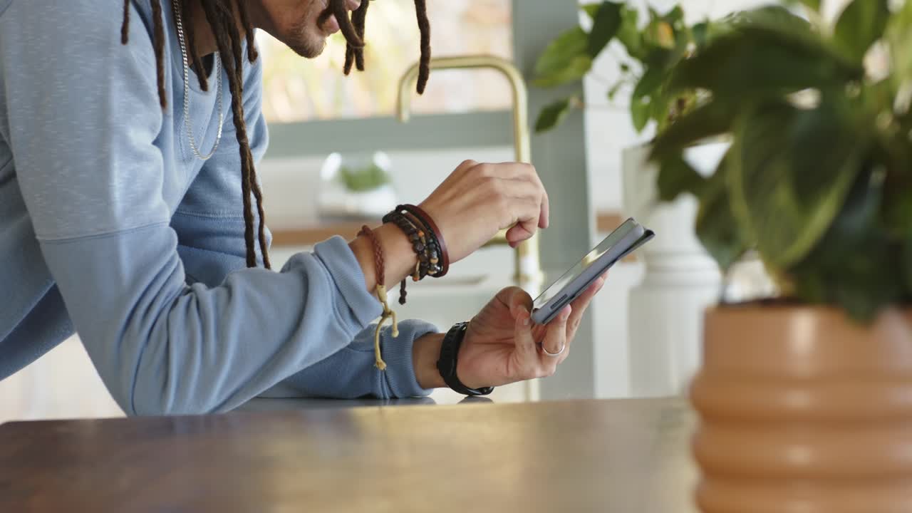 Leaning over countertop, adult male is tapping and swiping phone, browsing near faucet and plants