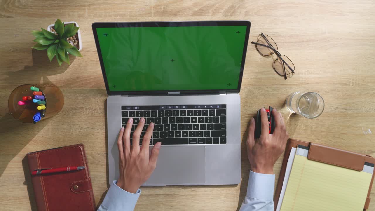 vista de arriba hacia abajo de una computadora portátil con pantalla verde simulada pantalla chromakey en un escritorio de oficina de madera al lado de la libreta con bolígrafos, gafas y un vaso de agua.
