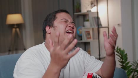 frustrated disappointed asian fat man watches tv while sitting on a couch at home