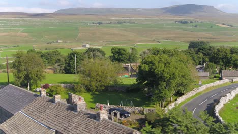 drone footage flying low over a dry stone wall sided country road and behind a roof top and trees showing the yorkshire countryside and fields with pen y ghent mountain in the distance