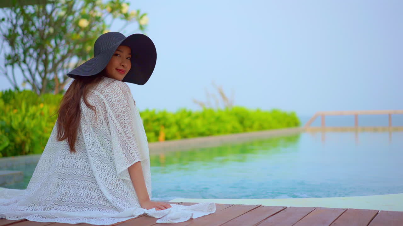 A woman dressed in a lacy cover-up and huge floppy hat sits on the deck of a resort pool looking out at the ocean, turns to look at the camera