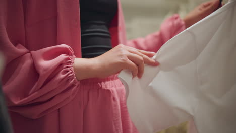 lower body view of a lady wearing an unbuttoned pink top thoughtfully evaluating a white polo shirt, bringing it close to her for inspection, the background shows a blur view of other clothes