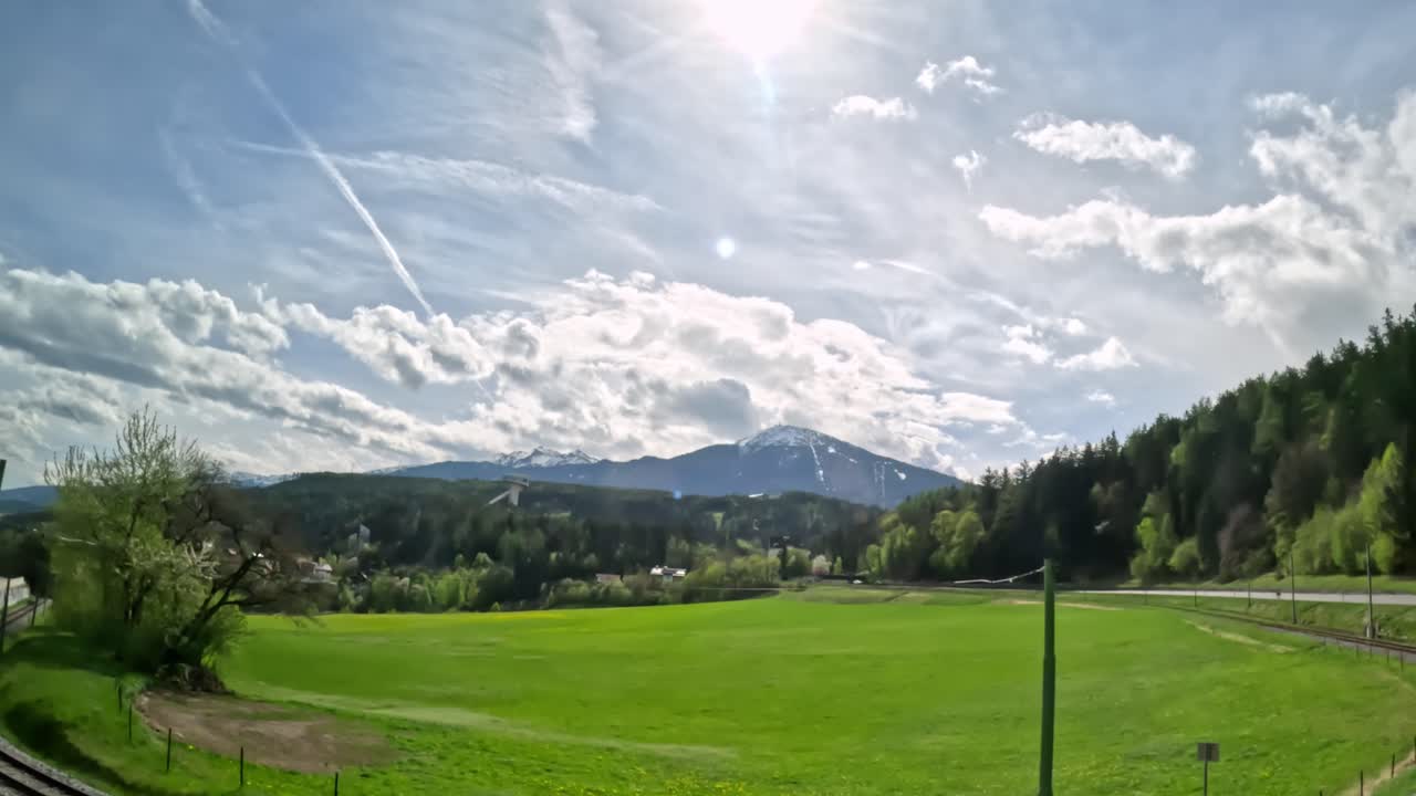 Sweeping panoramic extreme wide shot shows a green alpine valley near Innsbruck, Austria, framed by forested hills, distant snow-capped peaks, and a sky filled with large, dramatic springtime clouds