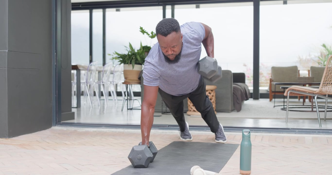 African American man in tee starting plank dumbbell-rows patio mat resting and drinking to hydrate