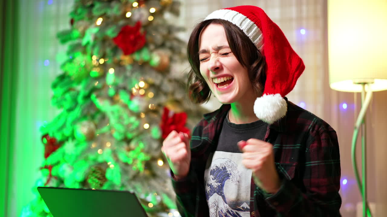 Woman in Santa cap looks at her laptop focused. Girl rejoices clapping hands receiving god news. Christmas tree at backdrop.