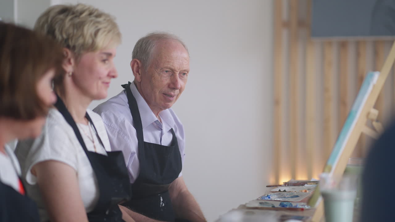 vista lateral de una persona mayor feliz sonriendo mientras dibuja como actividad recreativa o terapia en clase de pintura junto con el grupo de mujeres y hombres jubilados