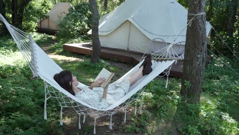 girl with dark hair in white country dress reads a book in a hammock (view 1)