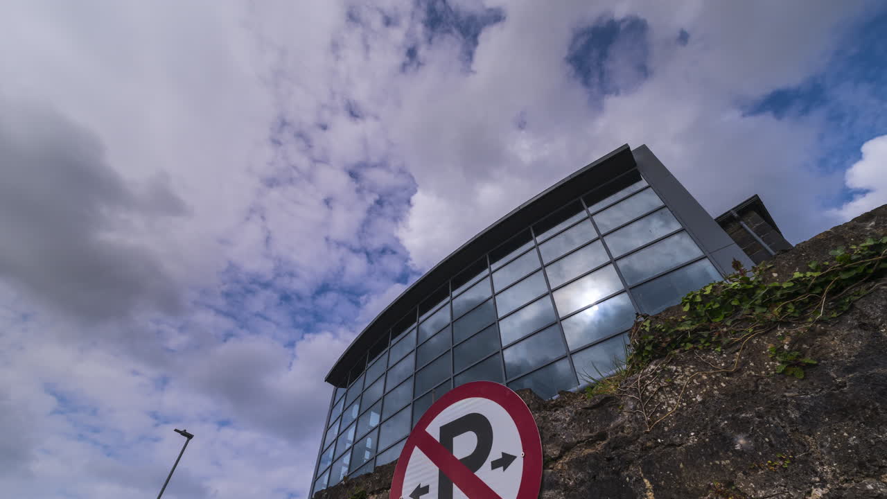 Timelapse of Carrick on Shannon modern glass building with stonewall and parking sign in the foreground in county Leitrim with fast moving clouds during the day in Ireland