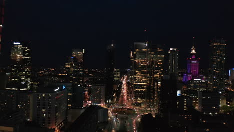 Forwards-fly-above-night-city.-Heavy-traffic-on-roads-and-intersection.-Illuminated-streets-and-tall-commercial-buildings-around.-Warsaw,-Poland