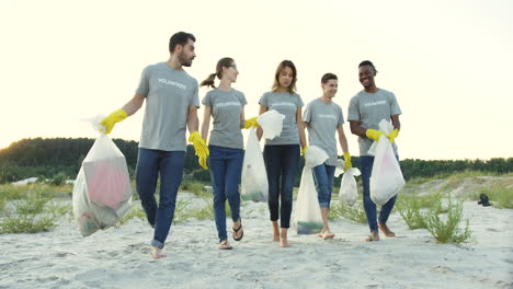 Gruppe-Junger-Multiethnischer-Menschen,-Die-Mit-Plastiktüten-Voller-Müll-Am-Strand-Spazieren-Gehen