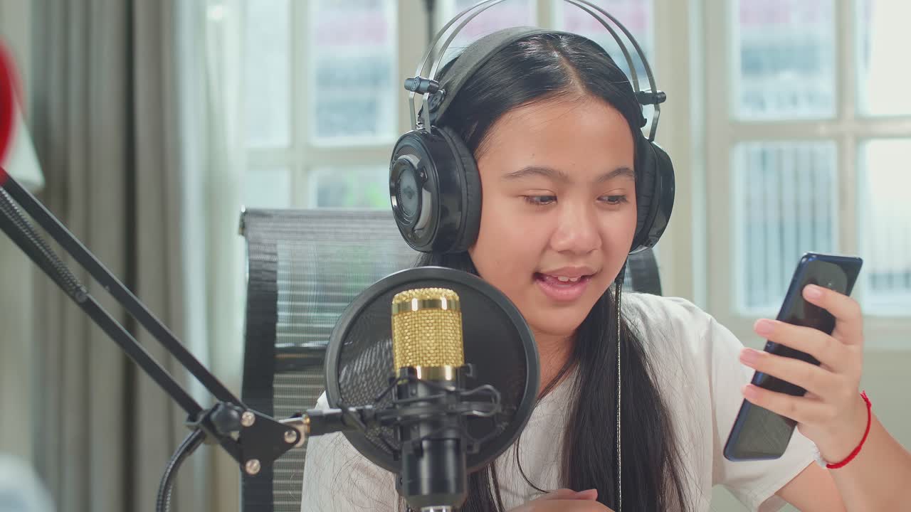 Over Shoulder View, Asian Kid Girl Holding Phone And Wearing Headphones Singing Into Microphone While Recording Podcast With Boy Host In Studio