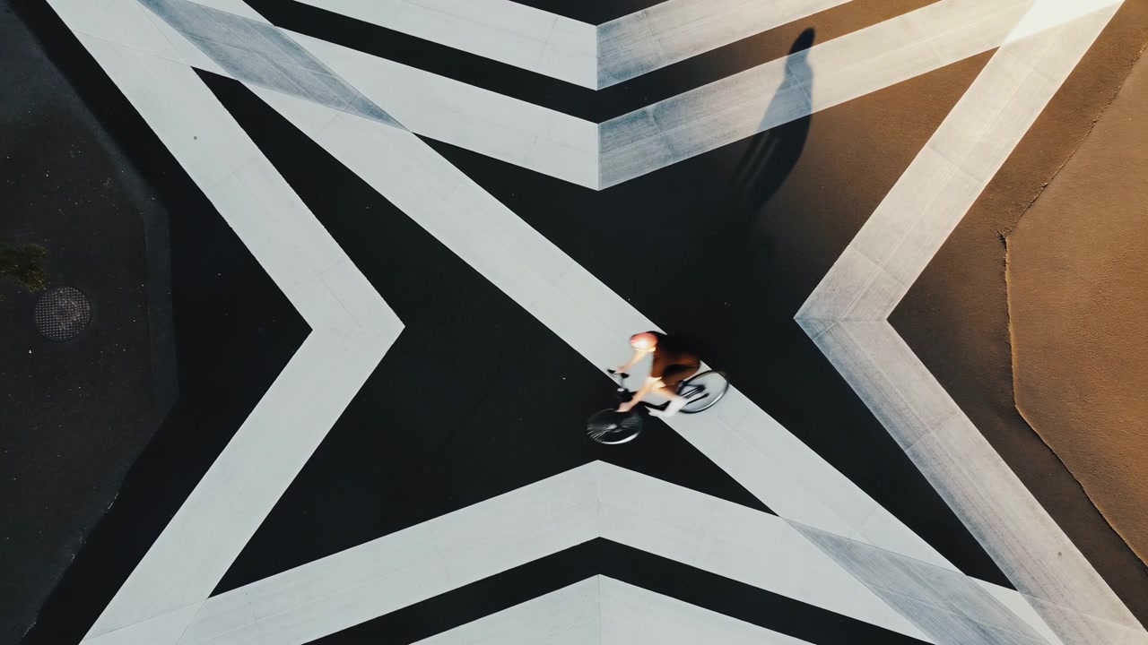 Vista dall'alto di un ciclista che pedala su una pavimentazione geometrica audace con forme a stella nere, bianche e beige, creando un forte contrasto e una texture grafica.