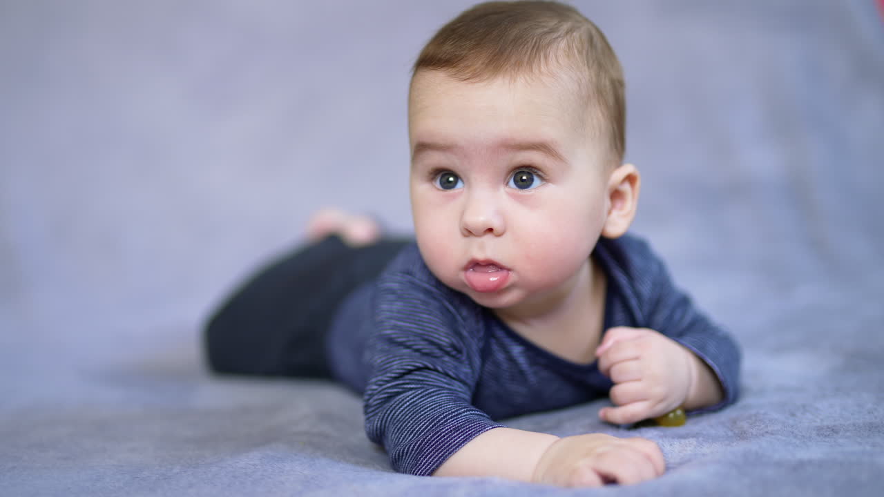 Lovely baby boy lies on belly looking up with surprise. Cute kid starts to move his limbs actively. Close up. Blurred grey backdrop.