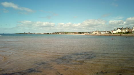 elie beach and harbour viewed from the beach cafe with children paddleboarding, sun shinning and beautiful blue sky