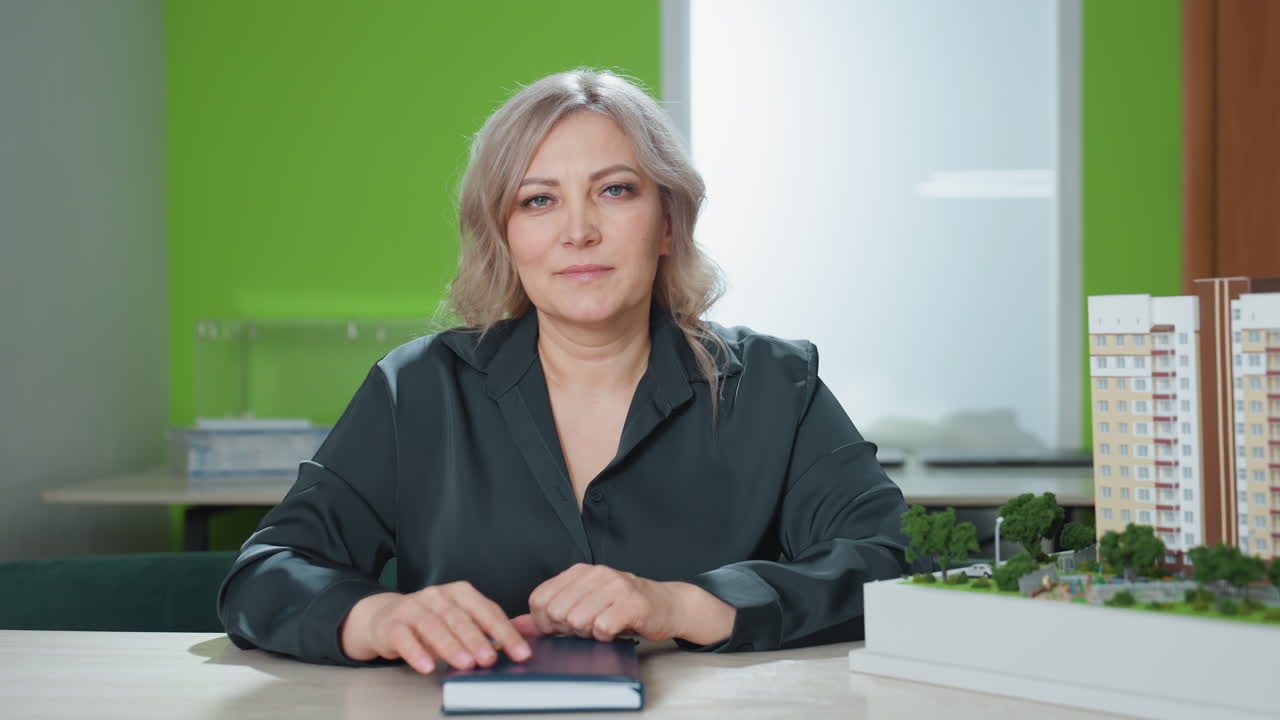 portrait view of young woman seated at desk closing book with poised gesture in modern office setting with green wall, building model, and organized workspace evoking elegance and professionalism