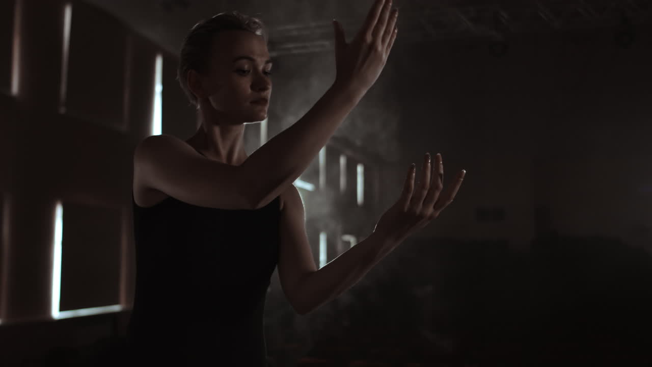 Prima ballet company in a dark dress on a dark theater stage rehearsing in the smoke performs dance moves in slow motion.