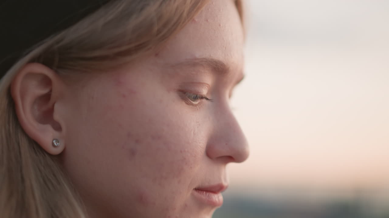 contemplative caucasian woman profile sunset, side profile in pastel dusk with freckled skin and stud earring, soft rim light on cheek and subtle highlight in eye, rooftop horizon blurred into