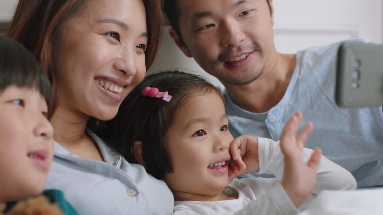 행복한 아시아 가족이 침대에서 스마트폰을 사용하여 비디오 채팅을 하고 있습니다. 엄마와 아빠는 아이들과 함께 손을 흔들며 휴대전화로 친구들과 채팅하고 있습니다. 온라인 커뮤니케이션 4k 영상을 즐기고 있습니다.