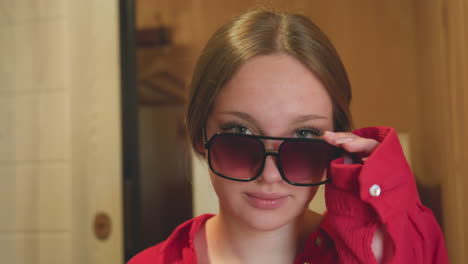 close up of young woman indoors wearing large sunglasses and red shirt while slightly lowering glasses to reveal eyes with calm, confident expression in softly lit wooden interior environment