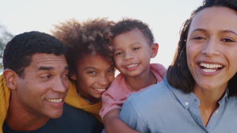 african american parents piggybacking their kids in the garden, looking to camera and laughing, close up