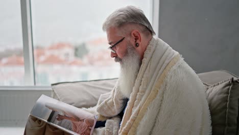 Side-view-of-a-man-with-gray-hair-with-a-full-beard-and-wearing-glasses-rapped-in-a-white-woolen-blanket-looks-through-a-magazine-while-sitting-on-the-sofa-in-a-modern-apartment-overlooking-the-sea