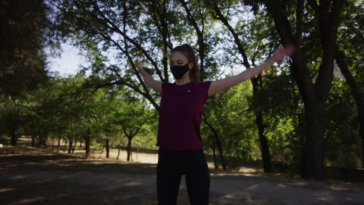 Young woman doing arm circles in a park using a face mask, medium shot