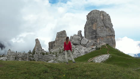 female hiker standing on alpine meadow, contemplating iconic cinque torri rock formations rising against dramatic dolomites landscape during summer mountain exploration