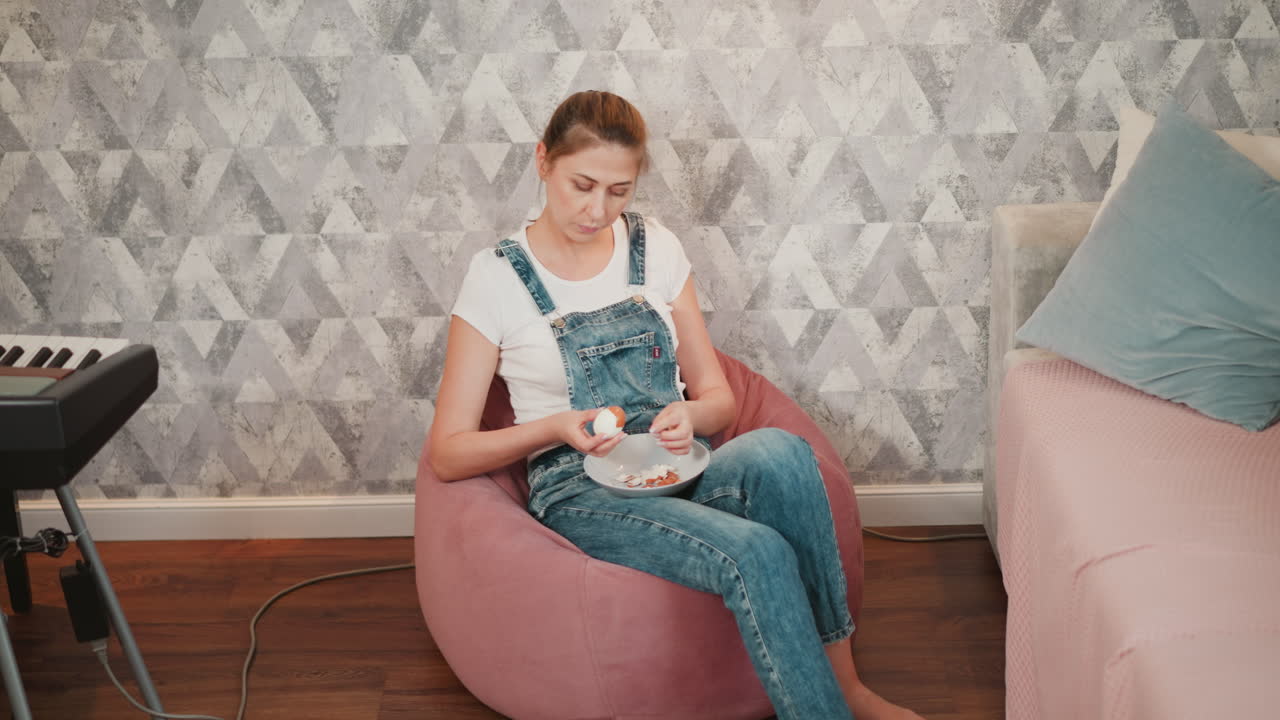 Young woman in denim overalls seated on pink bean bag peeling boiled egg into bowl, surrounded by cozy interior including partial view of keyboard and pastel couch